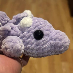Puede incluir: Un peque&ntilde;o juguete de peluche tejido a ganchillo, suave y de color lavanda claro. La criatura presenta una cabeza redondeada, un ojo de pl&aacute;stico negro liso y peque&ntilde;os detalles blancos y difusos en la parte superior de la cabeza y el lomo.