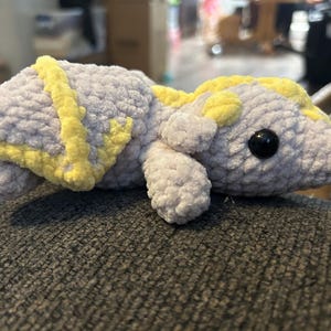 Puede incluir: Un peluche con forma de drag&oacute;n, hecho de hilo suave de color gris claro con detalles en amarillo brillante en las alas y el lomo. La criatura presenta una cabeza redondeada, extremidades peque&ntilde;as y un par de ojos de pl&aacute;stico negro brillante.