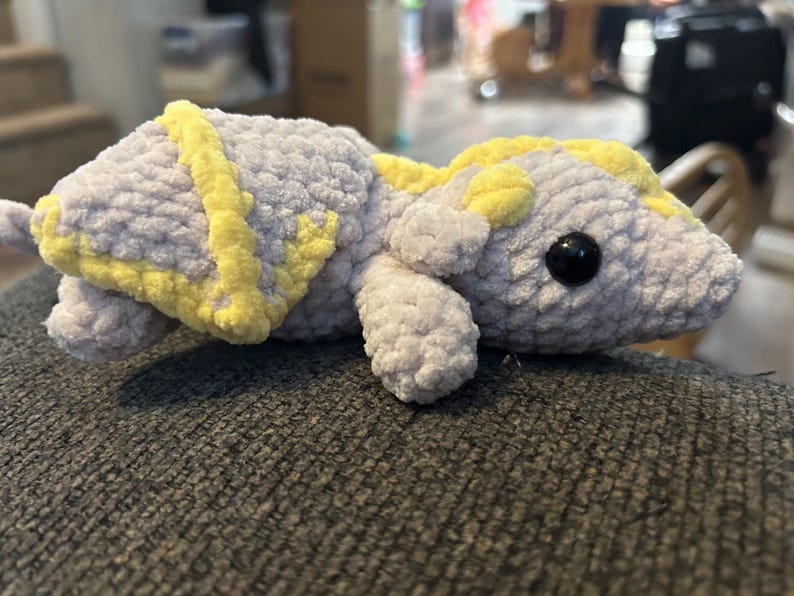 Puede incluir: Un peluche con forma de drag&oacute;n, hecho de hilo suave de color gris claro con detalles en amarillo brillante en las alas y el lomo. La criatura presenta una cabeza redondeada, extremidades peque&ntilde;as y un par de ojos de pl&aacute;stico negro brillante.