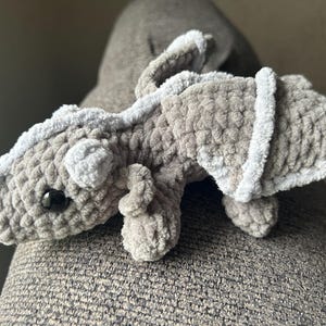 Puede incluir: Un drag&oacute;n de peluche hecho de hilo gris suave y texturizado. La criatura presenta una columna vertebral gris claro, alas peque&ntilde;as con bordes gris claro y un solo ojo de pl&aacute;stico negro visible. El cuerpo tiene una cabeza redondeada.