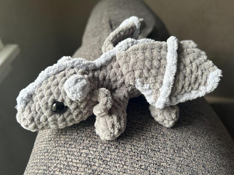 Puede incluir: Un drag&oacute;n de peluche hecho de hilo gris suave y texturizado. La criatura presenta una columna vertebral gris claro, alas peque&ntilde;as con bordes gris claro y un solo ojo de pl&aacute;stico negro visible. El cuerpo tiene una cabeza redondeada.