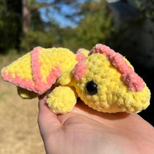 Puede incluir: Una peque&ntilde;a tortuga de ganchillo hecha a mano con un cuerpo amarillo brillante y detalles en rosa suave en el caparaz&oacute;n y la cabeza. La tortuga tiene una superficie de hilo texturizada y redondeada, una cola peque&ntilde;a y un ojo de seguridad negro.
