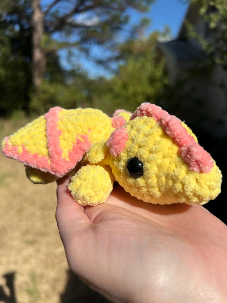 Puede incluir: Una peque&ntilde;a tortuga de ganchillo hecha a mano con un cuerpo amarillo brillante y detalles en rosa suave en el caparaz&oacute;n y la cabeza. La tortuga tiene una superficie de hilo texturizada y redondeada, una cola peque&ntilde;a y un ojo de seguridad negro.