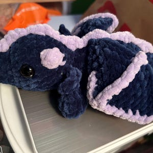 Puede incluir: Un drag&oacute;n de peluche hecho con hilo azul marino oscuro y lavanda claro. La criatura presenta una cabeza redondeada con un ojo negro, una espalda con crestas y dos alas grandes delineadas en lavanda. La textura es suave y esponjosa.