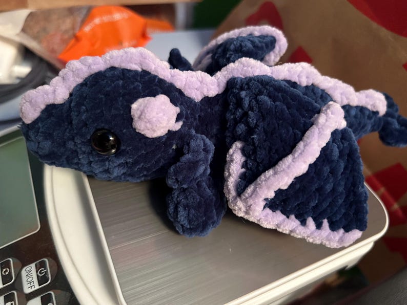 Puede incluir: Un drag&oacute;n de peluche hecho con hilo azul marino oscuro y lavanda claro. La criatura presenta una cabeza redondeada con un ojo negro, una espalda con crestas y dos alas grandes delineadas en lavanda. La textura es suave y esponjosa.