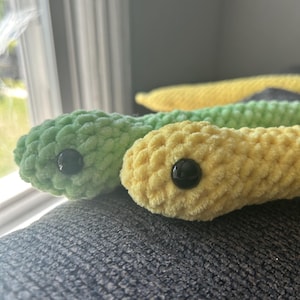 Puede incluir: Dos juguetes de serpiente de crochet, uno verde y otro amarillo, con ojos de botón negros.