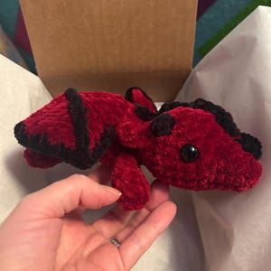 Puede incluir: Un drag&oacute;n de peluche hecho de hilo rojo oscuro suave con detalles de hilo negro en las alas y la cabeza. La criatura presenta dos peque&ntilde;os cuernos negros y ojos de pl&aacute;stico negro redondos y lisos, con una apariencia texturizada y difusa.