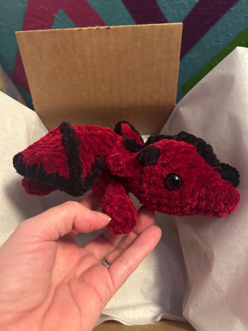 Puede incluir: Un drag&oacute;n de peluche hecho de hilo rojo oscuro suave con detalles de hilo negro en las alas y la cabeza. La criatura presenta dos peque&ntilde;os cuernos negros y ojos de pl&aacute;stico negro redondos y lisos, con una apariencia texturizada y difusa.