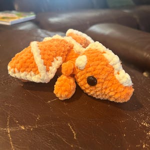 Puede incluir: Un peluche tejido a ganchillo con forma de lagarto o drag&oacute;n, elaborado con hilo naranja suave y detalles en hilo blanco a lo largo del lomo y la cabeza. La criatura presenta un hocico redondeado, extremidades peque&ntilde;as y un ojo de pl&aacute;stico negro.