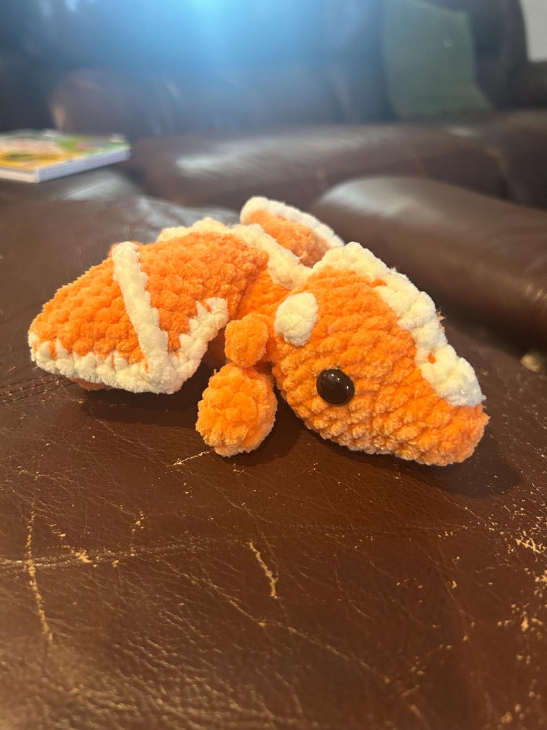 Puede incluir: Un peluche tejido a ganchillo con forma de lagarto o drag&oacute;n, elaborado con hilo naranja suave y detalles en hilo blanco a lo largo del lomo y la cabeza. La criatura presenta un hocico redondeado, extremidades peque&ntilde;as y un ojo de pl&aacute;stico negro.