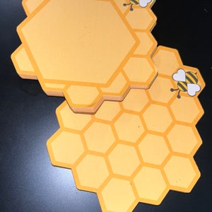 Puede incluir: Un conjunto de posavasos amarillos en forma de panal con un gr&aacute;fico de abeja de dibujos animados. Los posavasos tienen un centro amarillo claro con un borde amarillo m&aacute;s oscuro. El gr&aacute;fico de la abeja es negro y amarillo con alas blancas.