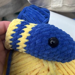 May include: A blue and yellow crocheted stuffed animal with a black button eye. The animal is made with fuzzy yarn and is sitting in a white tote bag.