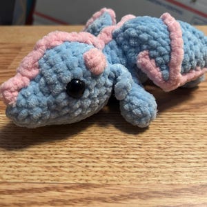 Puede incluir: Un peque&ntilde;o drag&oacute;n de peluche tejido a mano en ganchillo, hecho de hilo azul claro suave con detalles en hilo rosa claro a lo largo de la espalda, la cabeza y las alas. La criatura presenta un ojo de seguridad de pl&aacute;stico negro y textura.