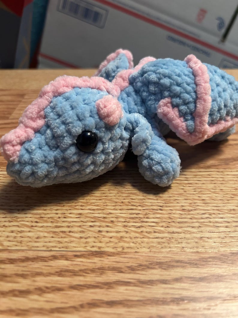 Puede incluir: Un peque&ntilde;o drag&oacute;n de peluche tejido a mano en ganchillo, hecho de hilo azul claro suave con detalles en hilo rosa claro a lo largo de la espalda, la cabeza y las alas. La criatura presenta un ojo de seguridad de pl&aacute;stico negro y textura.