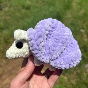 Puede incluir: Un juguete de caracol de ganchillo de felpa con una cabeza y cuerpo de color crema suave y una concha grande y texturizada de color lavanda. El caracol tiene un ojo de plástico negro, liso y redondo en el lateral, hecho de material de hilo difuso.