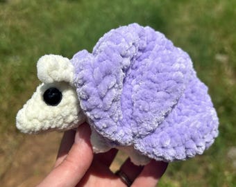 Puede incluir: Un juguete de caracol de ganchillo de felpa con una cabeza y cuerpo de color crema suave y una concha grande y texturizada de color lavanda. El caracol tiene un ojo de plástico negro, liso y redondo en el lateral, hecho de material de hilo difuso.