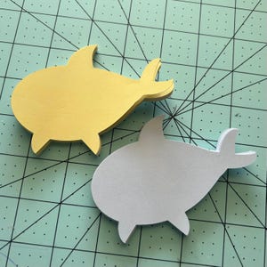 May include: Two paper shark cutouts, one yellow and one light gray, on a green cutting mat.