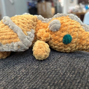 Puede incluir: Un drag&oacute;n de peluche hecho de hilo suave y texturizado en tonos amarillo dorado y gris claro. La criatura presenta una cabeza redondeada con un ojo circular de color verde azulado oscuro, orejas peque&ntilde;as y un dise&ntilde;o de lomo con crestas grises.