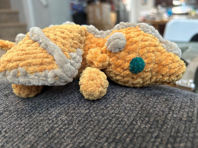 Puede incluir: Un drag&oacute;n de peluche hecho de hilo suave y texturizado en tonos amarillo dorado y gris claro. La criatura presenta una cabeza redondeada con un ojo circular de color verde azulado oscuro, orejas peque&ntilde;as y un dise&ntilde;o de lomo con crestas grises.
