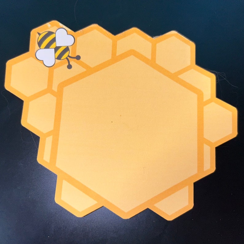 Puede incluir: Una pegatina amarilla con forma de panal de abeja con una ilustraci&oacute;n de abeja de dibujos animados. La abeja tiene rayas negras y amarillas con alas en forma de coraz&oacute;n. La pegatina es de color amarillo claro con un contorno amarillo m&aacute;s oscuro.