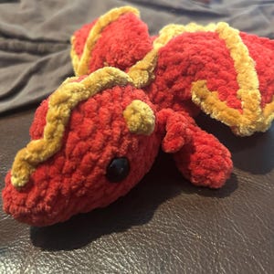Puede incluir: Un drag&oacute;n de peluche hecho de hilo rojo suave con detalles en amarillo dorado a lo largo de la espalda y las alas. La criatura presenta una cabeza redondeada, orejas peque&ntilde;as y un solo ojo de pl&aacute;stico negro visible. La textura es afelpada.