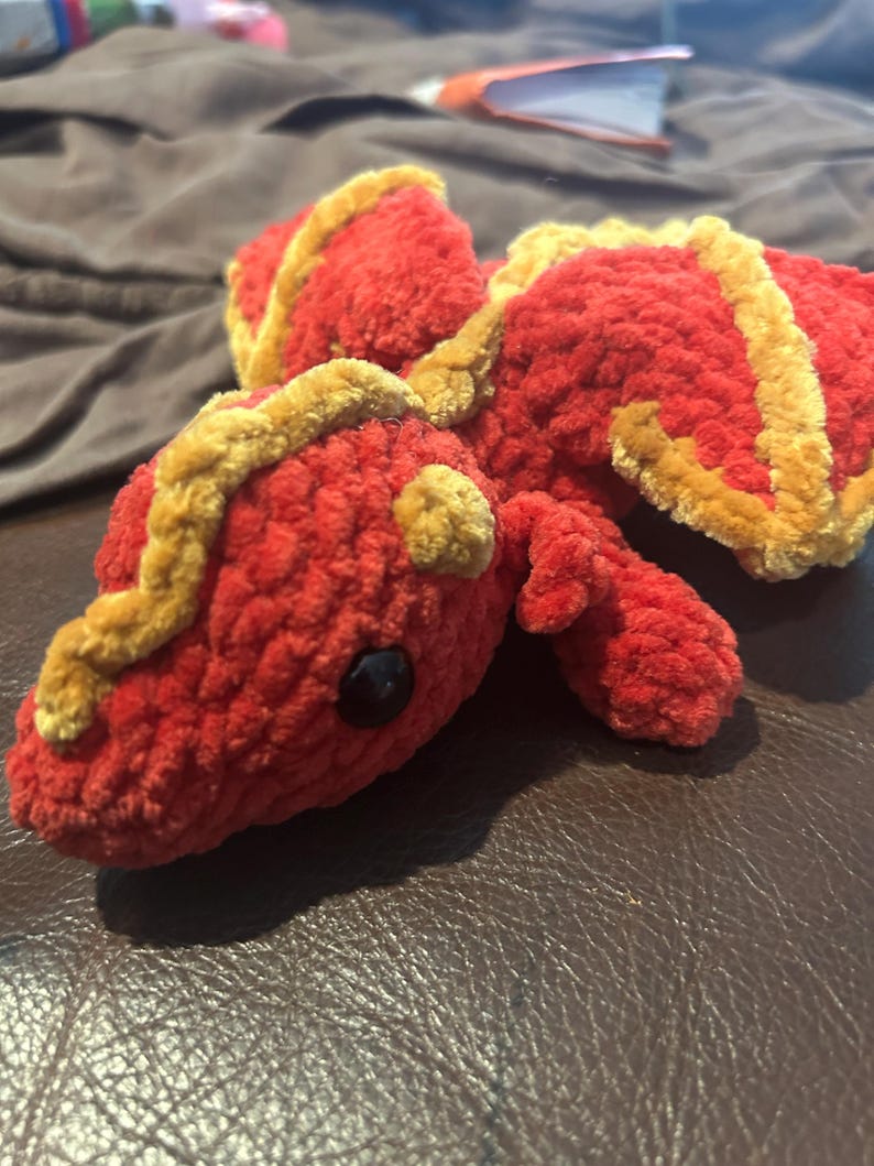 Puede incluir: Un drag&oacute;n de peluche hecho de hilo rojo suave con detalles en amarillo dorado a lo largo de la espalda y las alas. La criatura presenta una cabeza redondeada, orejas peque&ntilde;as y un solo ojo de pl&aacute;stico negro visible. La textura es afelpada.