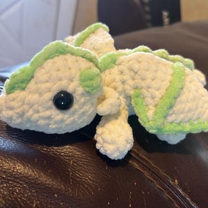 Puede incluir: Un peluche tejido a mano en forma de drag&oacute;n, elaborado con hilo blanco suave y detalles en verde claro a lo largo de la columna, las alas y la cabeza. La criatura presenta un &uacute;nico ojo de seguridad de pl&aacute;stico negro visible y una textura.