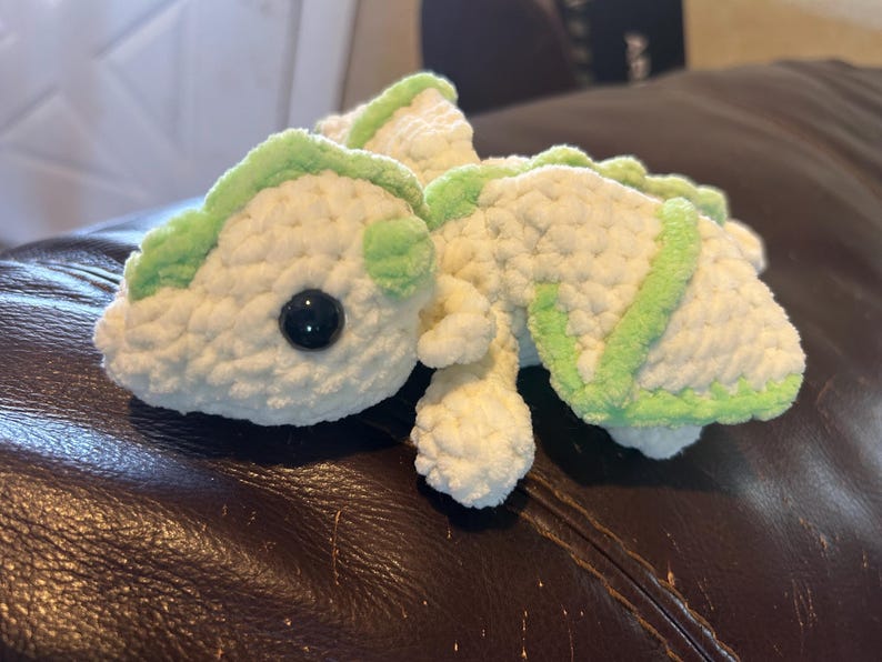 Puede incluir: Un peluche tejido a mano en forma de drag&oacute;n, elaborado con hilo blanco suave y detalles en verde claro a lo largo de la columna, las alas y la cabeza. La criatura presenta un &uacute;nico ojo de seguridad de pl&aacute;stico negro visible y una textura.