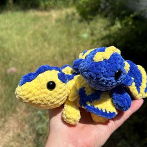 Puede incluir: Dos peque&ntilde;os peluches de ganchillo hechos a mano con una textura suave y difusa. Un peluche es principalmente amarillo con manchas azules, y el otro es principalmente azul con manchas amarillas. Ambos tienen ojos redondos negros y alas.