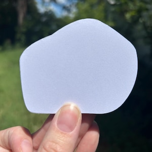 May include: A solid, light blue sticker with an irregular, rounded, organic shape. The sticker has a smooth, matte surface and is held up against a blurred outdoor background. The edges of the sticker are curved and asymmetrical, creating a unique silhouette.