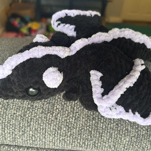 Puede incluir: Un drag&oacute;n de peluche hecho de hilo negro suave con detalles en lavanda claro a lo largo de las alas, la espalda y la cabeza. La criatura presenta un hocico redondeado, un solo ojo oscuro brillante visible y alas texturizadas.