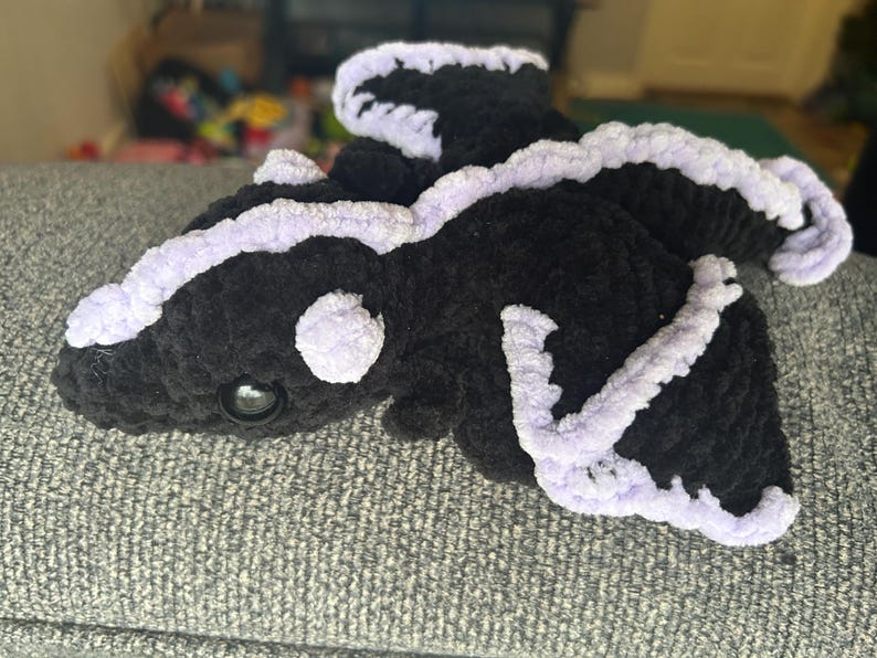 Puede incluir: Un drag&oacute;n de peluche hecho de hilo negro suave con detalles en lavanda claro a lo largo de las alas, la espalda y la cabeza. La criatura presenta un hocico redondeado, un solo ojo oscuro brillante visible y alas texturizadas.