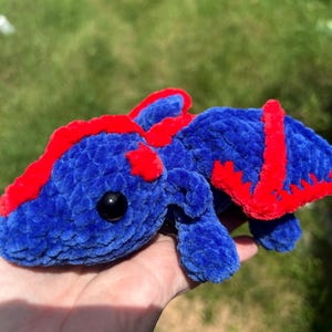 Puede incluir: Un drag&oacute;n de peluche hecho de hilo azul suave con acentos de color rojo vibrante a lo largo de la columna y las alas. La criatura presenta una cabeza redondeada, extremidades peque&ntilde;as y un par de alas, todo con una textura afelpada.