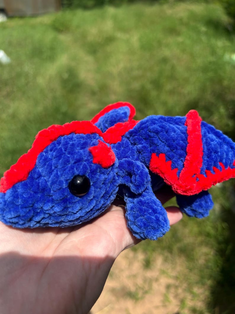 Puede incluir: Un drag&oacute;n de peluche hecho de hilo azul suave con acentos de color rojo vibrante a lo largo de la columna y las alas. La criatura presenta una cabeza redondeada, extremidades peque&ntilde;as y un par de alas, todo con una textura afelpada.