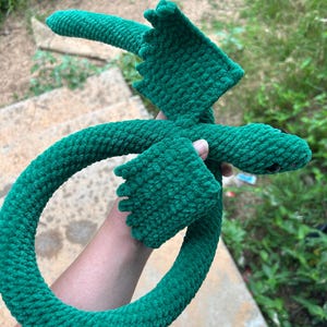 May include: A green crocheted dragon plush toy featuring a long, curved tail, two textured wings with scalloped edges, and a small head with dark, round eyes. The soft, yarn-based creature is crafted with a consistent stitch pattern throughout its body.