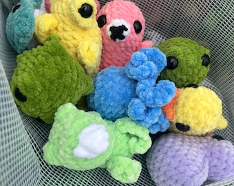 Puede incluir: Una colección de pequeñas criaturas de ganchillo, suaves y afelpadas, en varios colores como amarillo, rosa, azul, verde y morado. Cada juguete tiene ojos redondos de plástico negro y una superficie de hilo texturizado y esponjoso.