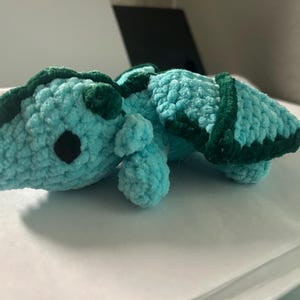 Puede incluir: Un peque&ntilde;o peluche de drag&oacute;n tejido a ganchillo a mano con hilo suave de color azul claro. La criatura presenta detalles en hilo verde oscuro a lo largo de la espalda y las alas, con dos peque&ntilde;os ojos redondos de pl&aacute;stico negro en la cabeza.
