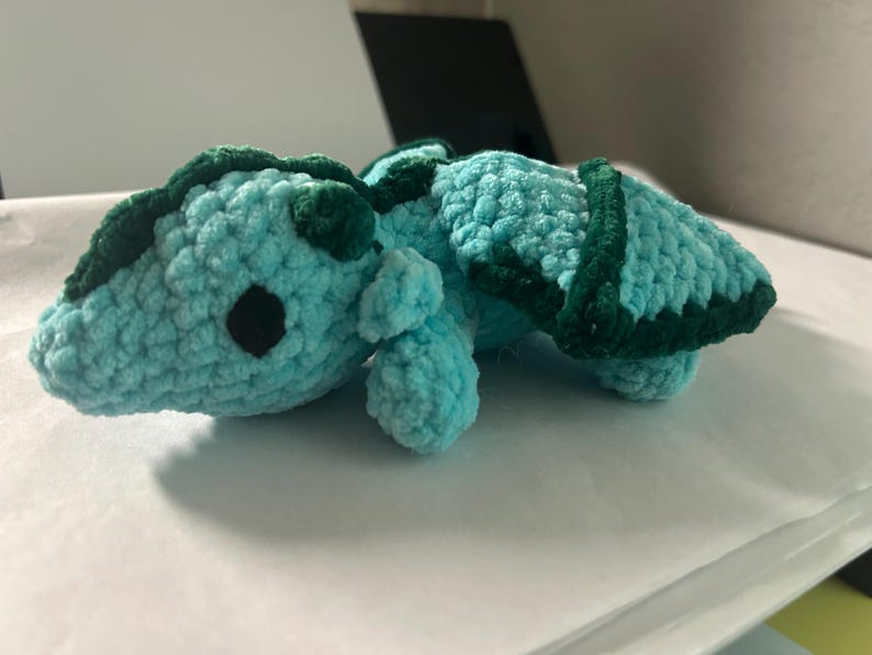 Puede incluir: Un peque&ntilde;o peluche de drag&oacute;n tejido a ganchillo a mano con hilo suave de color azul claro. La criatura presenta detalles en hilo verde oscuro a lo largo de la espalda y las alas, con dos peque&ntilde;os ojos redondos de pl&aacute;stico negro en la cabeza.