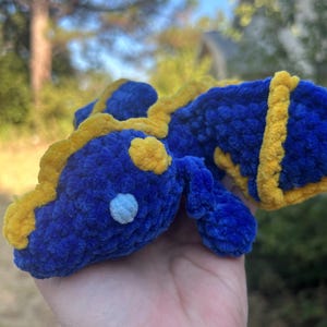 Puede incluir: Un peque&ntilde;o drag&oacute;n de peluche tejido a mano en ganchillo, hecho de hilo azul profundo suave con acentos de hilo amarillo brillante a lo largo de la espalda, la cabeza y las alas. La criatura presenta un detalle de ojo circular azul claro.