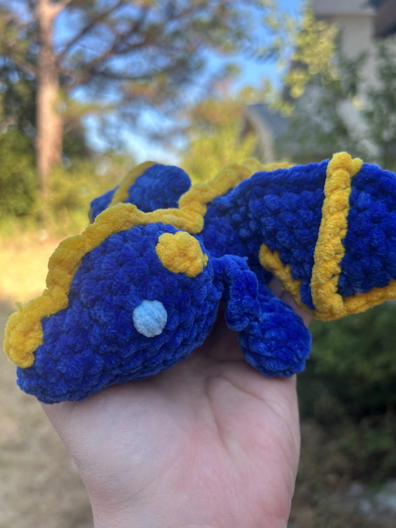 Puede incluir: Un peque&ntilde;o drag&oacute;n de peluche tejido a mano en ganchillo, hecho de hilo azul profundo suave con acentos de hilo amarillo brillante a lo largo de la espalda, la cabeza y las alas. La criatura presenta un detalle de ojo circular azul claro.