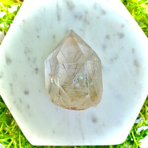 May include: A clear quartz crystal point with inclusions of golden rutile, creating a shimmering effect. The crystal is faceted and has a pointed tip. It is displayed on a white marble surface.