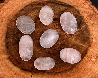 Puede incluir: Siete piedras ovaladas de cuarzo rosa con diseños de hojas talladas. Las piedras de color rosa pálido están dispuestas sobre una superficie de madera marrón oscuro y texturizada. Los grabados son detallados y visibles.