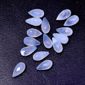 Puede incluir: Un conjunto de gemas en forma de l&aacute;grima de color azul claro, con superficies facetadas. Las gemas est&aacute;n dispuestas sobre un fondo oscuro y texturizado, mostrando su calidad transl&uacute;cida y sutiles variaciones de color. Ideal para la fabricaci&oacute;n de joyas o manualidades.