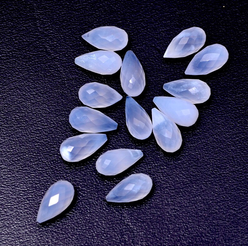 Puede incluir: Un conjunto de gemas en forma de l&aacute;grima de color azul claro, con superficies facetadas. Las gemas est&aacute;n dispuestas sobre un fondo oscuro y texturizado, mostrando su calidad transl&uacute;cida y sutiles variaciones de color. Ideal para la fabricaci&oacute;n de joyas o manualidades.