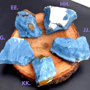 Könnte beinhalten: Fünf Stücke roher, hellblauer Opal-Edelsteine werden auf einer Holzscheibe präsentiert. Die Opale haben eine raue, natürliche Textur mit Farbvariationen und einigen braunen und weißen Einschlüssen. Die Buchstaben EE, GG, HH, JJ und KK sind sichtbar.