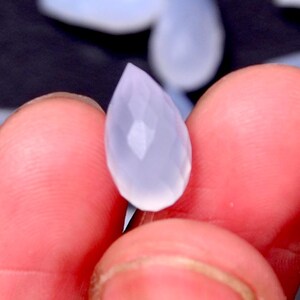 Puede incluir: Una gema facetada en forma de l&aacute;grima, de un tono azul p&aacute;lido y transl&uacute;cido. La gema se sostiene entre los dedos, mostrando sus superficies pulidas y su claridad interna. Las facetas de la gema capturan la luz, creando un brillo sutil.