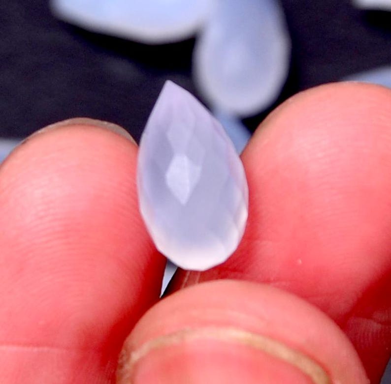 Puede incluir: Una gema facetada en forma de l&aacute;grima, de un tono azul p&aacute;lido y transl&uacute;cido. La gema se sostiene entre los dedos, mostrando sus superficies pulidas y su claridad interna. Las facetas de la gema capturan la luz, creando un brillo sutil.
