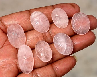 Puede incluir: Se muestran siete piedras preciosas ovaladas de color rosa claro con diseños de hojas talladas. Las piedras se sostienen en una mano, mostrando su calidad translúcida y sus intrincados detalles. Las piedras miden aproximadamente 2,5 cm de largo.