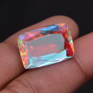 Op de afbeelding: Een grote, rechthoekige edelsteen met een regenboogirisatie. De edelsteen toont een spectrum aan kleuren, waaronder rood, blauw en groen, met een gepolijst, gefacetteerd oppervlak. De edelsteen wordt in een hand tegen een donkere achtergrond gehouden.