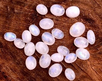 May include: A collection of oval-shaped moonstone cabochons. The stones are white with iridescent blue and purple flashes, scattered on a dark brown wooden surface. These polished gemstones are ideal for jewelry making.